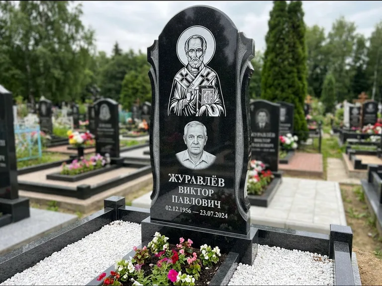 Памятник с гравировкой иконы: стела с образом в полный рост на обороте — гравировка образа Матроны Московской, низкие кусты спиреи в цвету, работа «Лебедева» в Омске